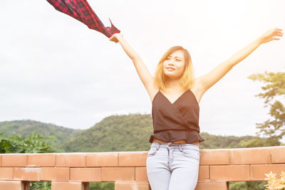 Smiling young woman with arms raised standing against sky
