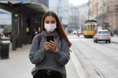 Young woman wearing mask using phone while standing outdoors