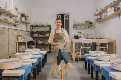 Confident craftsperson sitting on stool amidst pottery wheels at workplace