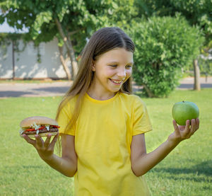 A teenage girl chooses between a burger and an apple outdoors in the park.