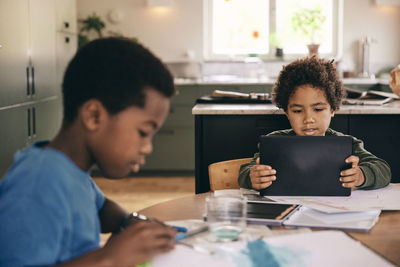 Boy using digital tablet while brother coloring in book at home