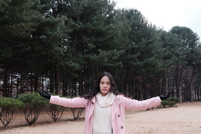 Portrait of young woman with arms outstretched standing against trees at park during snowfall