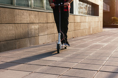 Unrecognizable crop male in checkered shirt riding modern electric scooter along pavement in city