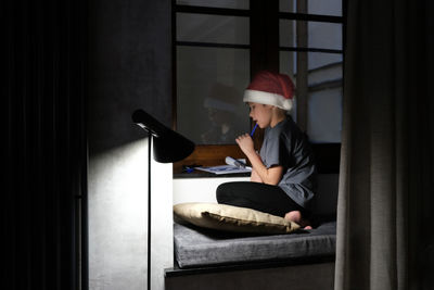 Boy wearing a santa claus hat writes a letter asking for a gift for christmas in a dark room.