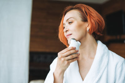 Woman in white bathrobe with red hair doing fasial massage with microcurrent facial massager