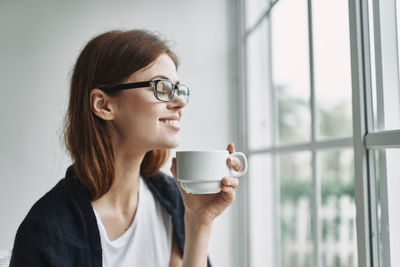 Smiling young woman holding coffee cup by window at home