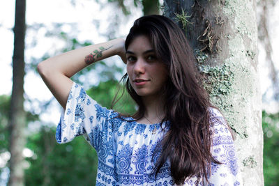 Portrait of beautiful young woman standing by tree trunk at park