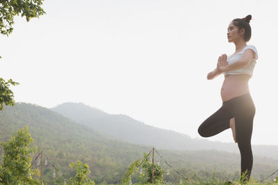 Pregnant woman exercising while standing on one leg against sky