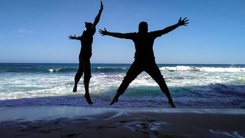 Silhouette friends jumping on shore at beach against sky