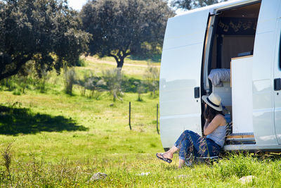 Woman sitting by van on grass against trees