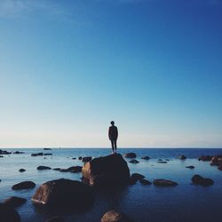 Man standing on rock by sea against clear blue sky