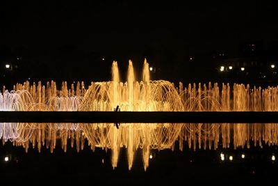 Fountain by lake against sky at night