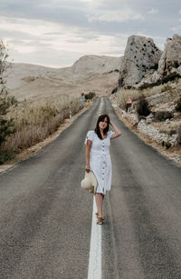 Front view of beautiful young woman in white dress standing on open road.