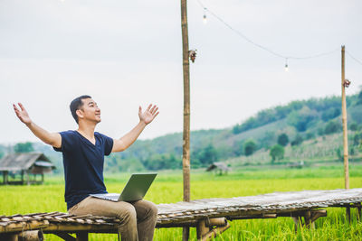 Man with arms outstretched using laptop while sitting on boardwalk at rice paddy