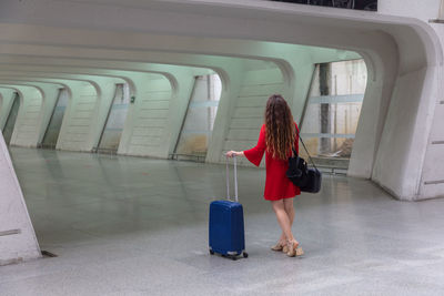 Back view of anonymous woman in red dress walking with suitcase in airport aisle