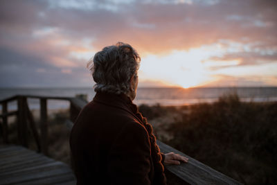 Rear view of man standing by railing against sea during sunset