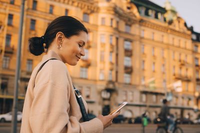 Fashionable smiling woman using mobile phone while walking against building in city