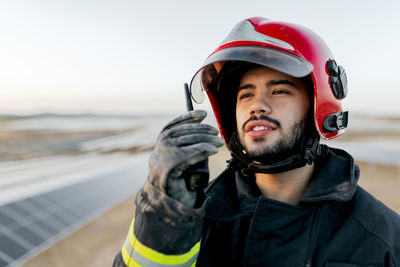 Positive young bearded male firefighter wearing helmet and protective worn gloves and clothes while holding walkie talkie in raised hand and peacefully looking away on blurred background in daytime