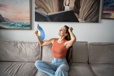 Smiling young woman fanning herself and waving while sitting on couch at home. heat and hot weather.