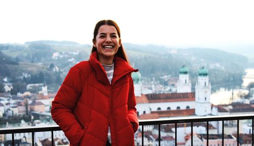 Portrait of smiling young woman in red jacket standing against cityscape