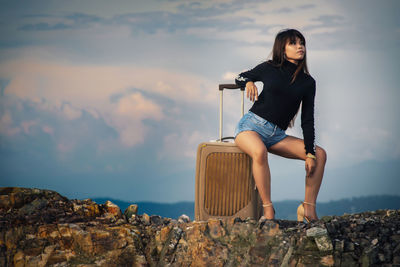 Young woman sitting on suitcase on cliff against cloudy sky