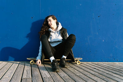 Teenage girl looking away while sitting on skateboard against wall