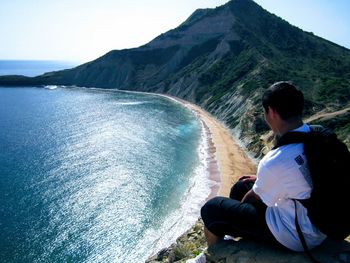Rear view of man sitting on mountain by sea against sky