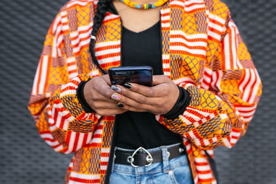 Cropped unrecognizable trendy young ethnic female in colorful clothes browsing on smartphone against gray wall on city street