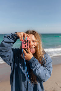 Cheerful young female in wet clothes taking photo on camera while standing with tongue out on sandy beach near waving sea