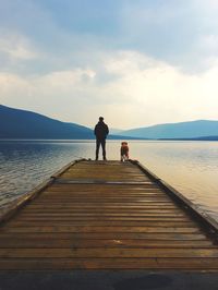 Rear view of man standing by dog on pier in lake and mountains against sky