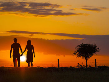 Silhouette couple walking on field against orange sky