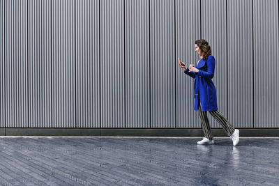 Full length side view of woman using phone while standing against wall