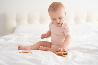 Baby girl in light pink bodysuit playing with wooden toys on white bedding on bed