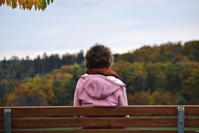 Rear view of woman sitting on bench against sky during autumn