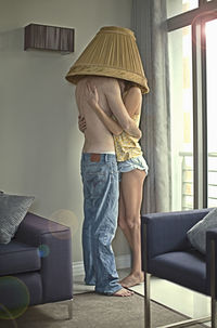 Young couple with lamp shade on head embracing each other while standing at home