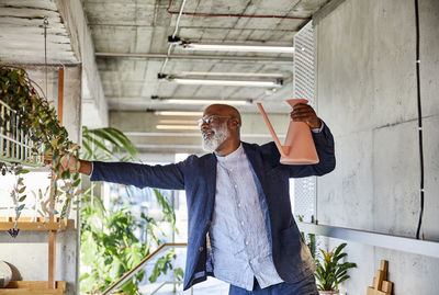 Mature man holding watering can looking at plant while standing at home