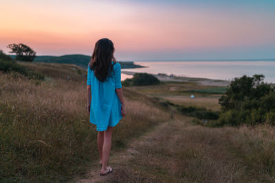 Rear view of woman walking on field against sky during sunset