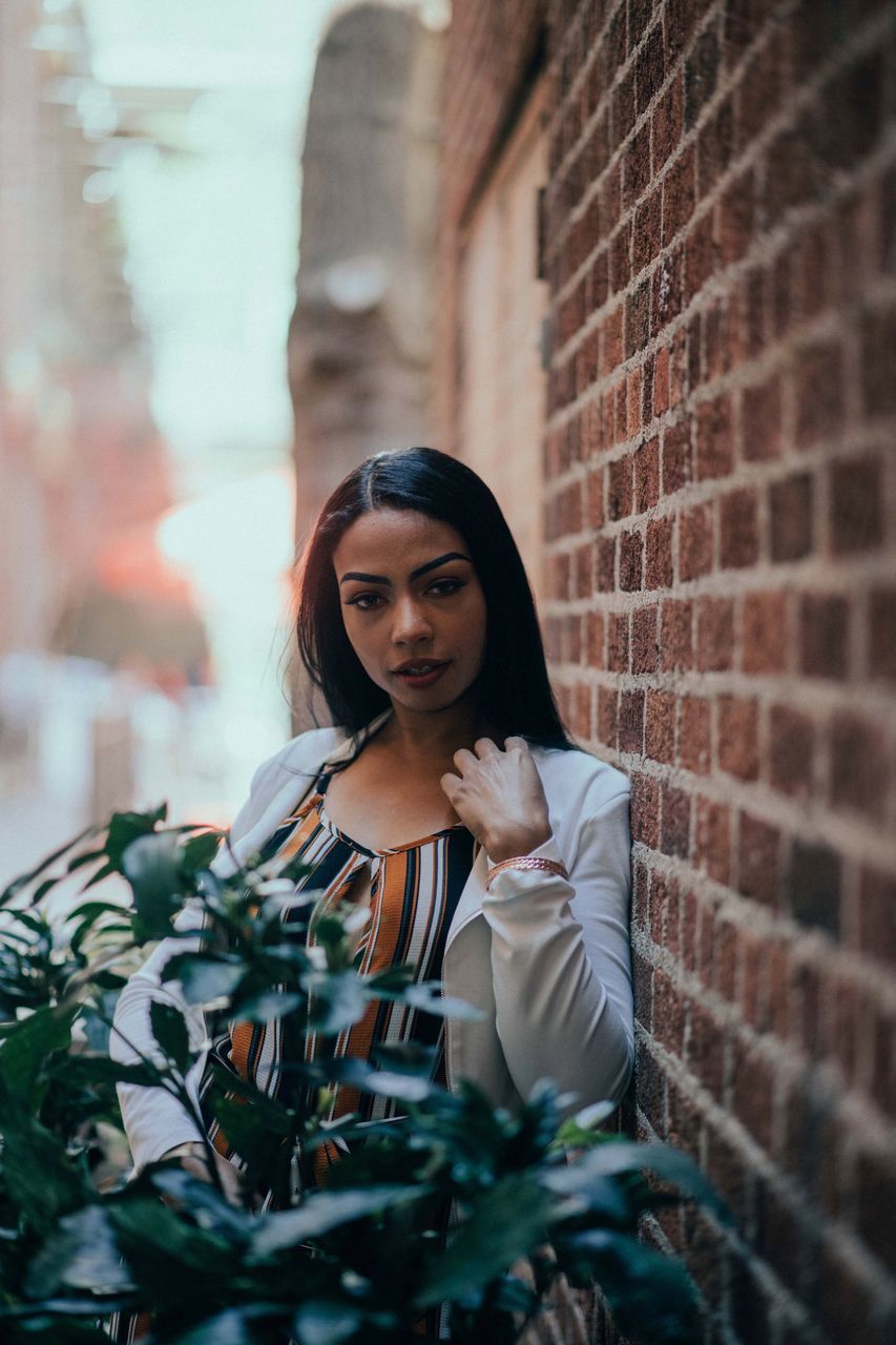 looking at camera, portrait, plant, one person, brick, brick wall, real people, young adult, front view, young women, lifestyles, selective focus, wall, flower, leisure activity, adult, women, flowering plant, standing, outdoors, hairstyle, beautiful woman