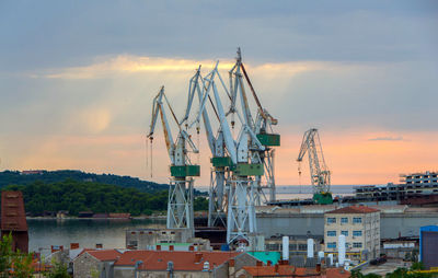 Cranes at harbor against sky during sunset