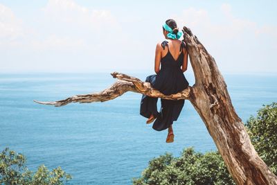 Full length rear view of woman sitting on branch by sea against sky