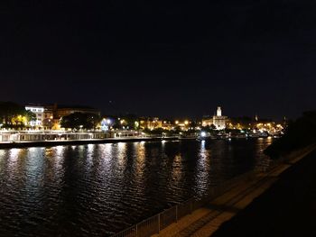 Illuminated buildings by river against sky at night