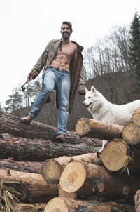 Portrait of young man standing with dog on logs of wood in forest against sky