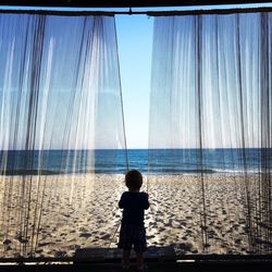 Rear view of boy holding curtains while standing in gazebo at beach