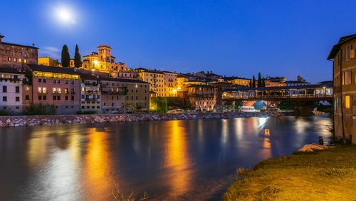 Illuminated buildings by river against sky at night