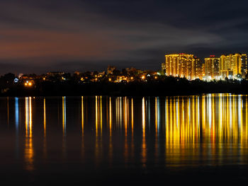Illuminated buildings by river against sky at night