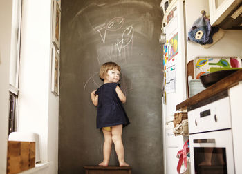 Rear view of girl writing on wall while standing on stool at home