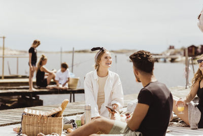 Smiling friends talking while sitting over jetty on sunny day during picnic