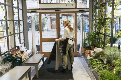 Full length of female owner holding cardboard while standing at pathway in store