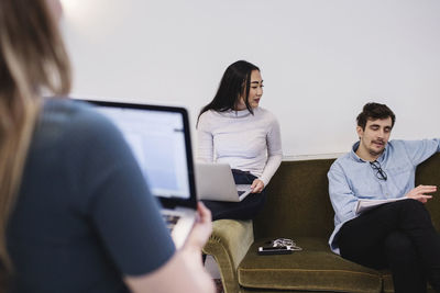 Young woman looking at male colleague sitting on sofa at office