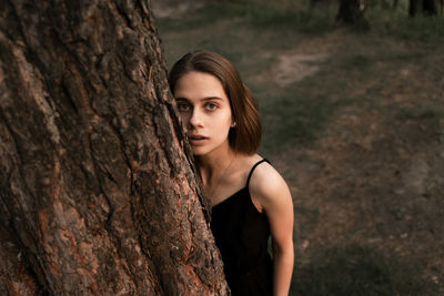 Portrait of young woman standing against tree trunk in forest